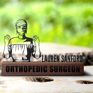 May include: A wooden plaque with the name "Lauren Sanford" and the title "Orthopedic Surgeon" in black text. A black silhouette of a female surgeon wearing a surgical mask and gown stands above the plaque.