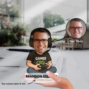 Puede incluir: Caricatura de jugador personalizada con una cara sonriente que lleva gafas y auriculares. La figura está sentada, sosteniendo un mando de juego y vistiendo una camiseta negra con la palabra "GAMER". Incluye una foto y espacio para nombre y texto personalizados.