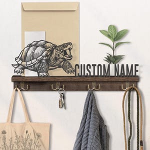 May include: A wooden wall shelf featuring a metal turtle silhouette and the text "CUSTOM NAME". The shelf has four hooks, holding a tote bag, keys, a scarf, and a leash. A small plant is also on the shelf.