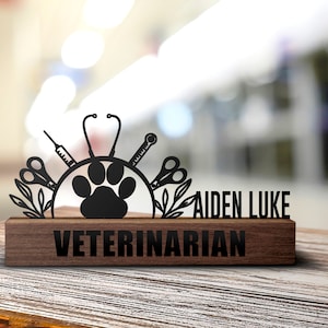 May include: Black metal sign with a paw print, stethoscope, scissors, and syringe. The sign reads "Aiden Luke Veterinarian" and is mounted on a wooden base.