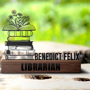 Puede incluir: Una placa de madera con el texto "Benedict Felix Librarian" y una silueta negra de una pila de libros con un diseño floral en la parte superior.