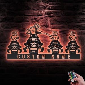 May include: A black metal silhouette of four snowmobilers with a custom nameplate. The snowmobilers are riding in a line, with the first snowmobiler leading the way. The snowmobilers are illuminated with a warm orange glow. The nameplate is illuminated with a warm orange glow. The nameplate says "CUSTOM NAME".