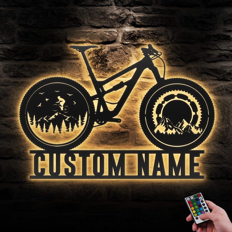 May include: Black metal mountain bike wall art with a backlit design. The bike silhouette features a rider, trees, and mountains within the wheels. The text "CUSTOM NAME" is below the bike. A remote control is in the lower right corner.