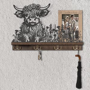 May include: A rustic wall-mounted key holder featuring a metal silhouette of a Highland cow with a floral crown. The key holder has four brass hooks, a small shelf, and a framed photo. An umbrella hangs from one of the hooks.