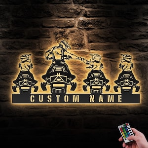 May include: A black metal silhouette of four people riding snowmobiles with the text "CUSTOM NAME" below. The silhouette is illuminated with warm white light. A remote control with multiple colored buttons is visible in the bottom right corner.