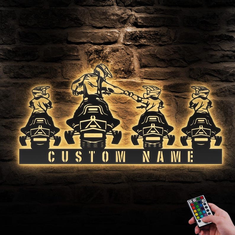 May include: A black metal silhouette of four people riding snowmobiles with the text "CUSTOM NAME" below. The silhouette is illuminated with warm white light. A remote control with multiple colored buttons is visible in the bottom right corner.