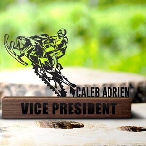 May include: A black and green metal sculpture of a person riding a snowmobile, with the text "CALEB ADRIEN" above a wooden base that reads "VICE PRESIDENT". The sculpture is set against a blurred green background.