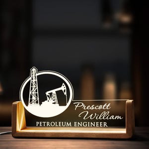 May include: A wooden desk nameplate with a white acrylic insert featuring an oil rig and pump jack design. The name "Prescott William" and the title "Petroleum Engineer" are also engraved on the acrylic insert.