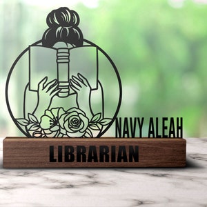 Puede incluir: Una silueta de metal negro de una persona leyendo un libro con flores y el texto "NAVY ALEAH LIBRARIAN" sobre una base de madera.