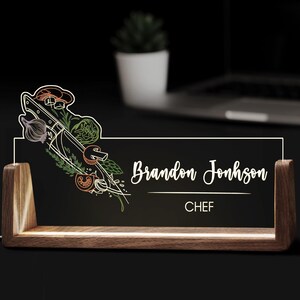 May include: A wooden desk nameplate with the name "Brandon Jonhson" and the title "CHEF" in white script. The nameplate features a colourful illustration of a chef's knife, vegetables, and herbs. The nameplate is illuminated with a soft glow.