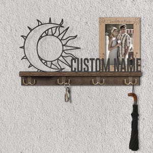 Puede incluir: Un llavero de madera marrón oscuro con cuatro ganchos de latón. Presenta un diseño de sol y luna de metal, las palabras "CUSTOM NAME" y una foto enmarcada. Un paraguas cuelga de uno de los ganchos.