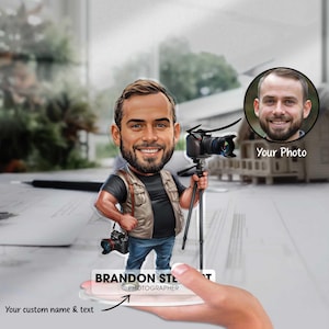 May include: A cartoon illustration of a person holding a camera and tripod. The person is wearing a blue shirt, khaki vest, and blue jeans. The text "BRANDON STEPHENS PHOTOGRAPHER" is on the base of the figurine.
