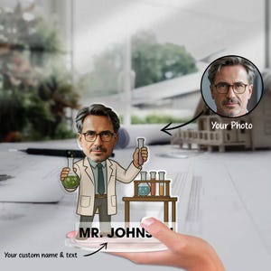 Può includere: Figura di scienziato personalizzata in acrilico. L'illustrazione in stile cartone animato raffigura un uomo con un camice da laboratorio, che tiene in mano becher e provette. La figura include il testo "MR. JOHNS" e uno spazio per una foto personalizzata.