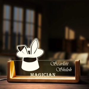 May include: A wooden base with a clear acrylic panel featuring a white silhouette of a magician's hat with a rabbit sticking out of it. The text "Scarlett Shiloh" and "MAGICIAN" are printed on the panel.
