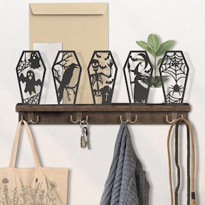 May include: A dark brown wooden shelf with five black coffin-shaped Halloween decorations. Each coffin features a different spooky design, including a ghost, raven, skeleton, witch, and spiderweb. The shelf has brass hooks below, with a tote bag, keys, and a sweater hanging.