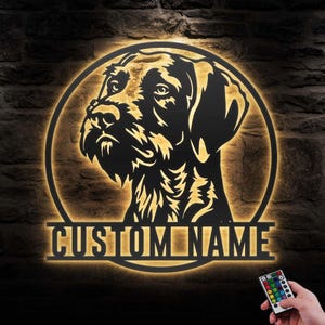 Könnte beinhalten: Ein schwarzes Metallschild mit einer Hunde-Kopf-Silhouette in einem kreisförmigen Rahmen, beleuchtet mit einem warmen Schein. Der Text "CUSTOM NAME" ist darunter. Eine Fernbedienung wird unten rechts gehalten.