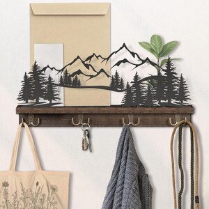 May include: A rustic wall-mounted shelf with a dark wood finish, featuring a black metal mountain and forest silhouette. The shelf includes four brass hooks for hanging items. A canvas tote bag, keys, and a scarf are hanging from the hooks.