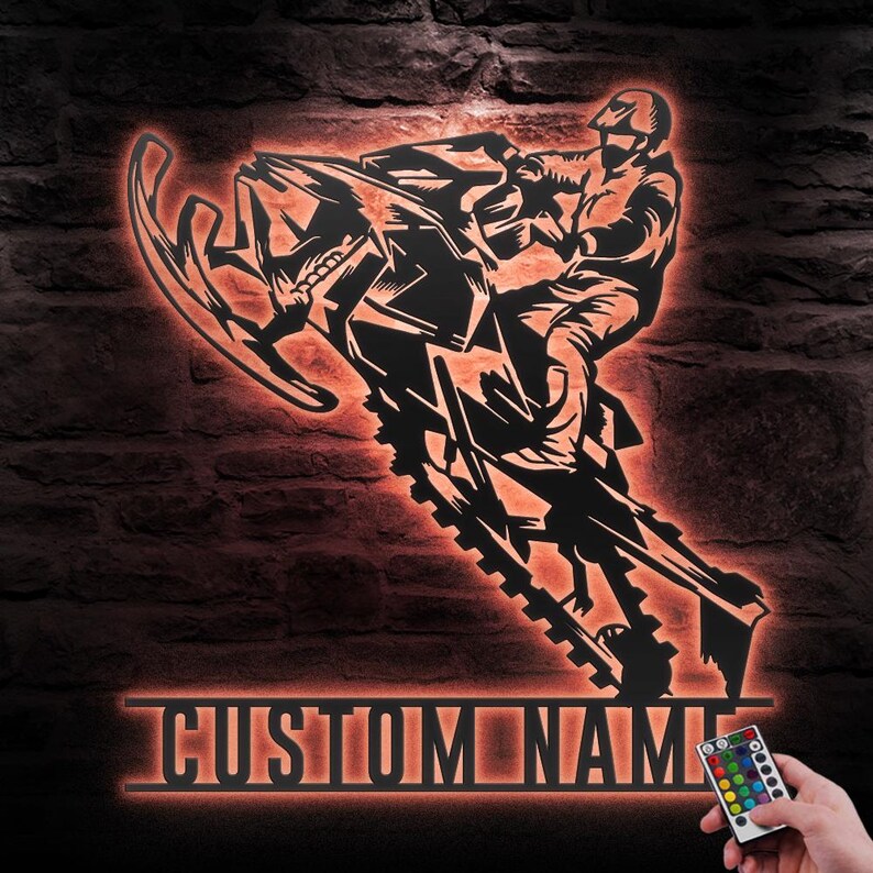 Puede incluir: Una silueta negra de metal de una persona conduciendo una moto de nieve con una placa de nombre personalizada debajo. La silueta est&aacute; iluminada con un brillo naranja c&aacute;lido. Un control remoto con m&uacute;ltiples luces de colores es visible en la esquina inferior derecha.