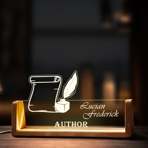 May include: A wooden desk lamp with a clear acrylic panel. The panel features a scroll, quill, and inkwell design, along with the name "Lucian Frederick" and the word "AUTHOR". The lamp emits a warm, inviting light.