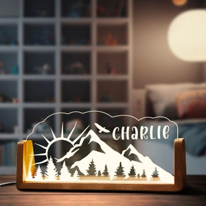 May include: A wooden night light with a clear acrylic insert featuring a mountain scene with a sun, trees, and a bird flying over the mountains. The name "Charlie" is engraved on the acrylic insert.