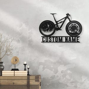 May include: Black metal mountain bike wall art with a custom name plate. The bike silhouette features detailed wheels with mountain and forest scenes. The text "CUSTOM NAME" is displayed below the bike.