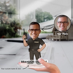 Puede incluir: Una caricatura personalizada de guardia de seguridad sosteniendo un walkie-talkie. La figura lleva un uniforme oscuro con "SECURITY" en el pecho y un cinturón. La base muestra el nombre "BRANDON STEWART" y "SECURITY". Se muestra una foto junto a la caricatura.