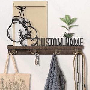 May include: A dark brown wooden wall shelf with four brass hooks, a decorative boxing glove silhouette, and the text "CUSTOM NAME". A tote bag, keys, a scarf, and a jump rope hang from the hooks. A small plant sits on the shelf.