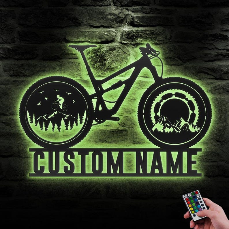 May include: Black metal mountain bike wall art with a green neon glow. The bike silhouette features detailed wheels with mountain and forest scenes. The text "CUSTOM NAME" is below the bike. A remote control is in the lower right corner.