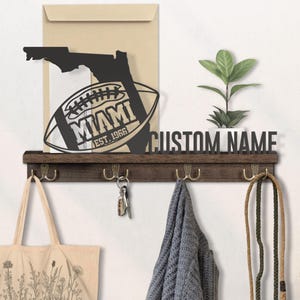 May include: A dark brown wooden wall-mounted shelf with four brass hooks. The shelf features a black metal cutout of the Florida state outline and a football with "MIAMI EST. 1966" text. A "CUSTOM NAME" sign is above the shelf.