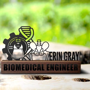 May include: A wooden desk nameplate with the words "BIOMEDICAL ENGINEER" in black. Above the nameplate is a black and green graphic of a gear, DNA strand, and skeletal elements. The name "ERIN GRAY" is also displayed.