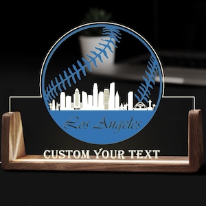 May include: A wooden desk lamp with a blue baseball design featuring the Los Angeles skyline. The lamp has a customizable text area and a warm glow. The baseball design is approximately 20 cm in diameter.