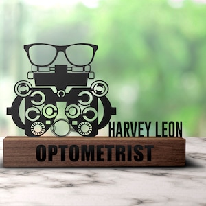 Könnte beinhalten: Schwarze Metallsilhouette eines Augenprüfdiagramms mit Brille oben, montiert auf einer Holzplakette mit dem Text "HARVEY LEON OPTIKER".