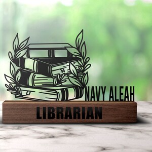Puede incluir: Corte de metal negro de libros apilados con enredaderas de hojas, montado en una placa de madera oscura con el texto "NAVY ALEAH LIBRARIAN".
