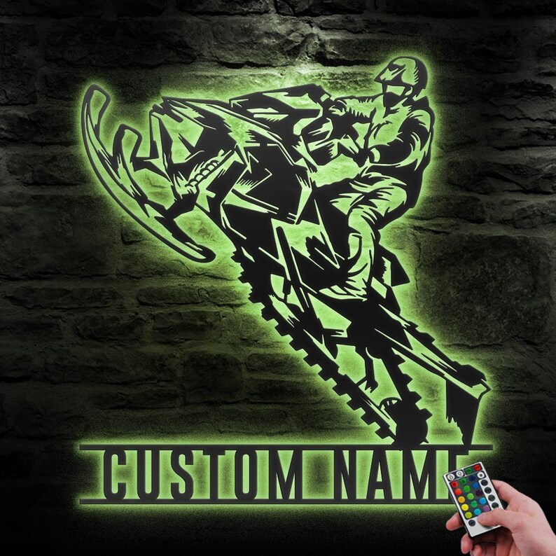 Puede incluir: Una silueta negra de metal de una persona conduciendo una moto de nieve con una placa de nombre personalizada debajo. La silueta est&aacute; iluminada en verde con un mando a distancia multicolor.
