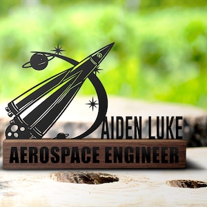 Op de afbeelding: Een zwart metalen raket met sterren en planeten, gemonteerd op een houten plaat met de tekst "Aiden Luke Aerospace Engineer".