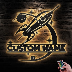 May include: A black metal silhouette of a rocket ship with a crescent moon and stars. The rocket ship has a custom name plate that reads "CUSTOM NAME". The silhouette is illuminated with warm white light.