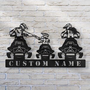 May include: Black metal silhouette of three people riding snowmobiles. The snowmobiles are side by side and the people are holding onto the handlebars. The snowmobiles have a custom name plate that says "CUSTOM NAME".