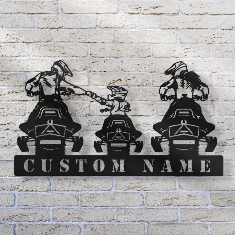 May include: Black metal silhouette of three people riding snowmobiles. The snowmobiles are side by side and the people are holding onto the handlebars. The snowmobiles have a custom name plate that says "CUSTOM NAME".