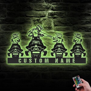 May include: A black metal silhouette of four snowmobilers with a custom nameplate. The snowmobilers are riding in a line, with the first snowmobiler leading the way. The snowmobilers are illuminated with a green light. The image includes a remote control with multiple colored buttons.