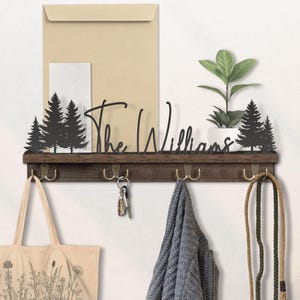 May include: A wooden wall-mounted shelf with hooks, featuring a black metal family name and silhouette trees. The shelf holds a plant, keys, and a tote bag. The hooks are brass-colored.