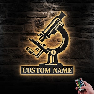 May include: A black metal silhouette of a microscope with a custom nameplate that reads "CUSTOM NAME". The silhouette is illuminated with warm white light and is mounted on a brick wall. A remote control with multiple colored buttons is visible in the bottom right corner of the image.