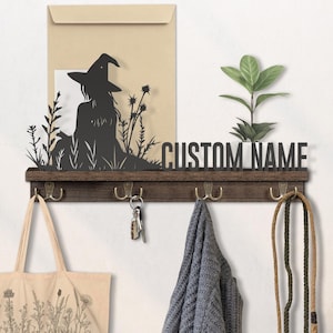May include: A dark brown wooden wall-mounted coat rack with four brass hooks. It features a black metal silhouette of a witch in a field, a space for a custom name, and a small plant. A tote bag, keys, a scarf, and a leash hang from the hooks.