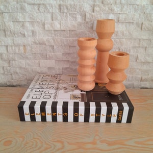 May include: Three wooden candle holders in different sizes and shapes. The candle holders are made of natural wood and have a light brown color. The candle holders are sitting on a black and white striped book with the title "Elements of Style" on the spine.
