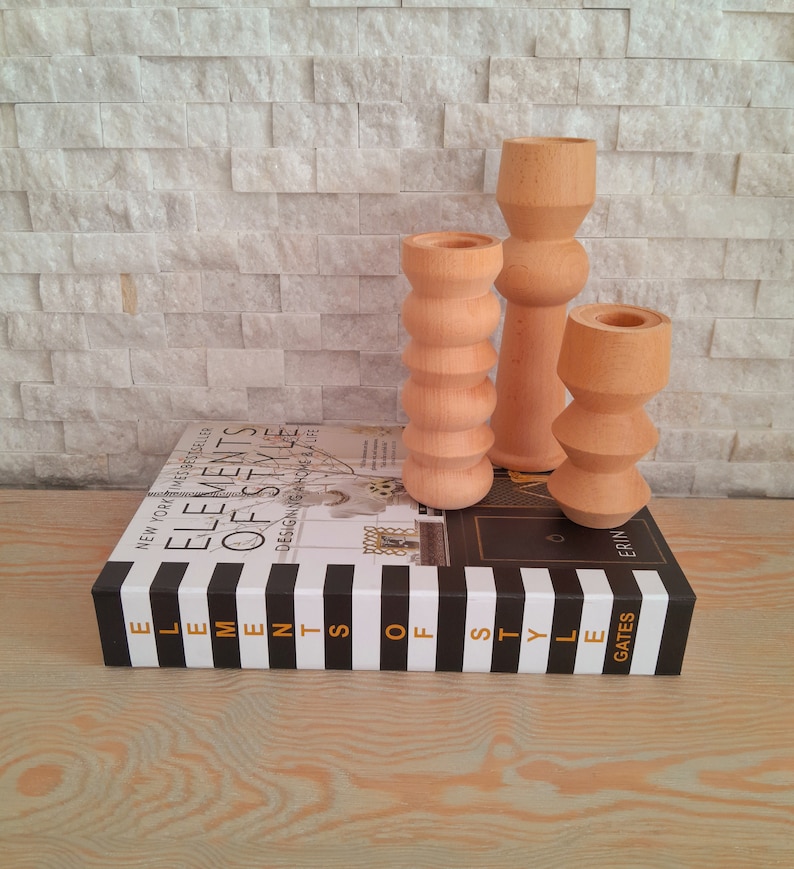 May include: Three wooden candle holders in different sizes and shapes. The candle holders are made of natural wood and have a light brown color. The candle holders are sitting on a black and white striped book with the title "Elements of Style" on the spine.