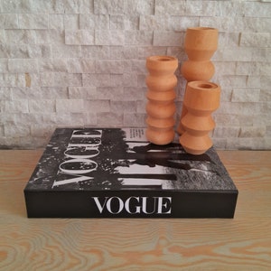 May include: Three wooden candle holders in different sizes, each with a unique, rounded, textured design. The candle holders are arranged on top of a black book with the word "VOGUE" printed on the cover.