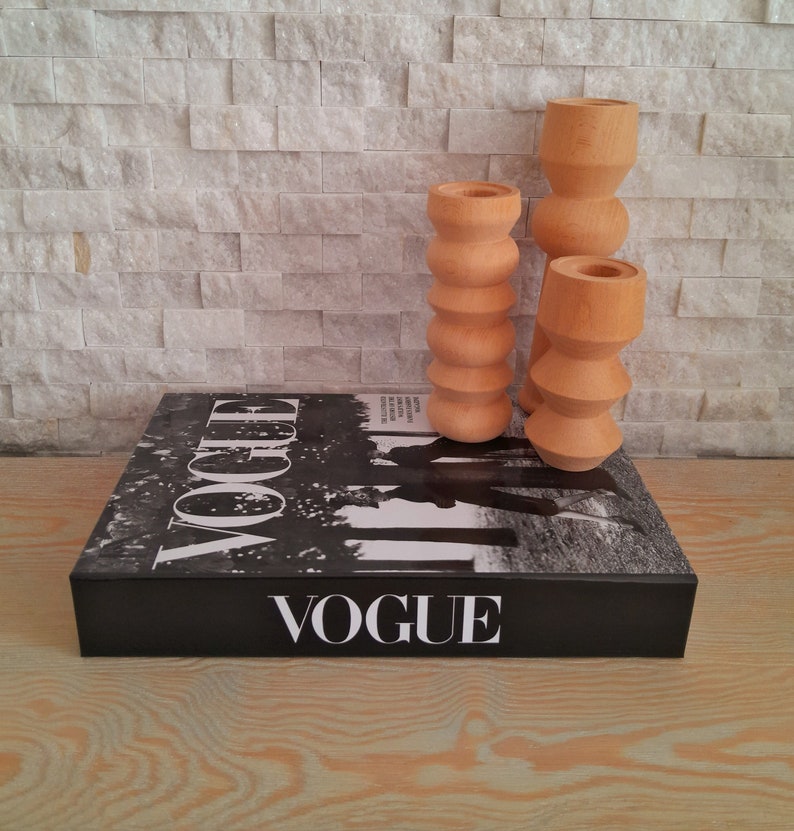 May include: Three wooden candle holders in different sizes, each with a unique, rounded, textured design. The candle holders are arranged on top of a black book with the word "VOGUE" printed on the cover.