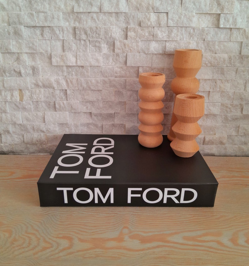 May include: A black book with the text "TOM FORD" printed in white on the cover. Three wooden candle holders in a light brown color are placed on top of the book.