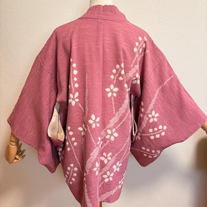 May include: A dusky pink kimono with white floral embroidery. The back of the garment is visible, showcasing the intricate design of white flowers and stems. The fabric has a textured appearance, and the sleeves are wide and flowing.