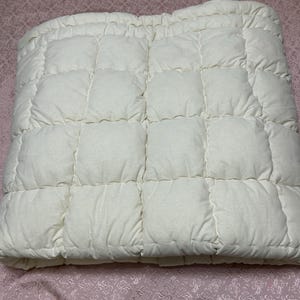 May include: A white quilted bed topper with a square pattern. The topper is folded in half and is lying on a pink patterned bedspread.
