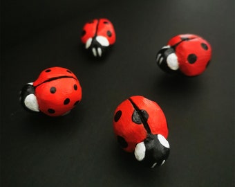 May include: Four hand-painted ladybugs, each with black spots and white accents, are arranged on a black surface. The ladybugs are made of a smooth, rounded material.